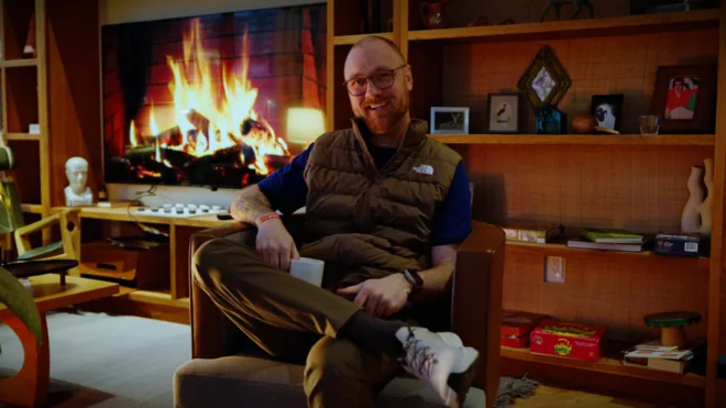 A still from the reactive video: Jake sits in an armchair with a mug, in front of a television playing a fireplace.