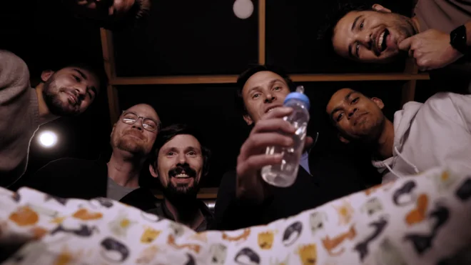 Six people crowded around, looking down into the camera from above a crib.