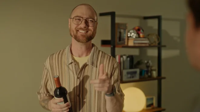 The host, holding a bottle of wine, mid-explanation to a guest.