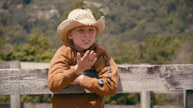A boy in a straw cowboy hat leaning on a paddock fence, watching.